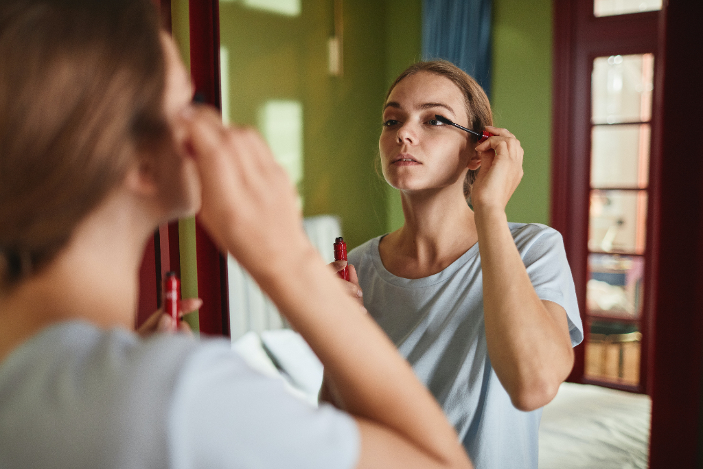 woman looking at a mirror and putting mascara on her eyes